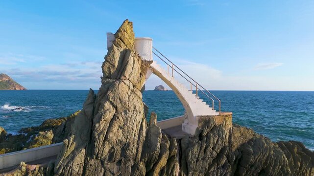 Aerial footage of El Clavadista in Mazatlan, Sinaloa, Mexico, capturing the rustic stairs, rocky cliff, and waves of the Pacific Ocean. Daring cliff divers, coastal beauty, and vibrant seaside.