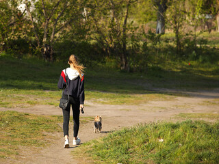A young girl walks in the park with a dog. A sporty blonde walks a pet in nature. View of a beautiful girl from the back. Relax in the fresh air with your loved one. Friendship between children