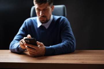 Man in sweater focused on smartphone at desk in dimly lit office environment during evening hours