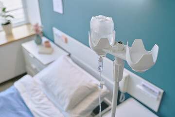 Empty hospital bed with intravenous drip stand positioned beside pillow in medical patient room, showing clean bedding and clinical equipment in healthcare facility setting
