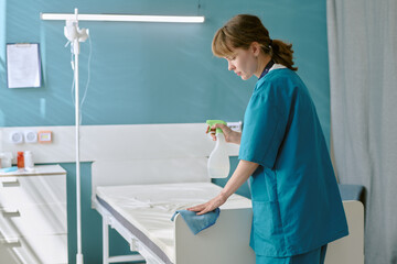 Fototapeta premium Caucasian young adult woman wearing medical scrubs cleaning hospital bed with spray bottle and cloth, standing in patient room, focusing on disinfecting surface for hygiene