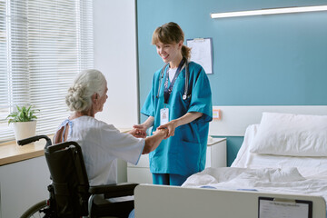 Obraz premium Caucasian senior woman with disability sitting in wheelchair holding hands with young adult Caucasian female nurse smiling in hospital room during medical care interaction