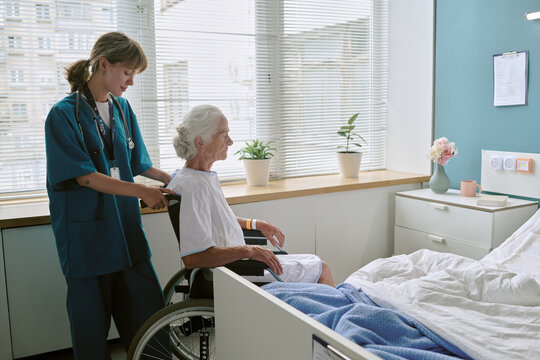 Young adult Caucasian female nurse assisting senior Caucasian woman sitting in wheelchair near hospital bed, nurse standing behind patient, medical care setting visible through window
