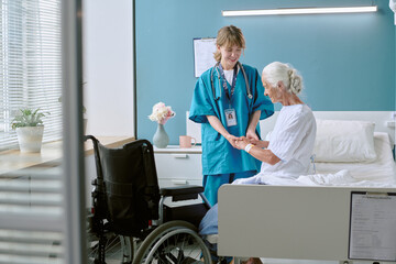 Caucasian female nurse assisting senior Caucasian woman sitting on hospital bed, holding hands and smiling, wheelchair positioned nearby in modern medical room