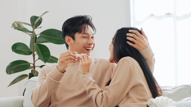 Young Asian couple sitting close on couch laughing joyfully while holding pregnancy test, concept of excitement, pregnancy and emotional bonding at home