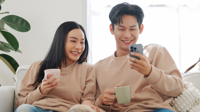Asian couple in sweaters sitting close on sofa smiling while looking at smartphone together, holding coffee mugs