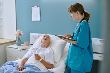 Senior Caucasian woman lying in hospital bed talking to young adult Caucasian female nurse standing beside bed, holding digital tablet and listening attentively during medical consultation