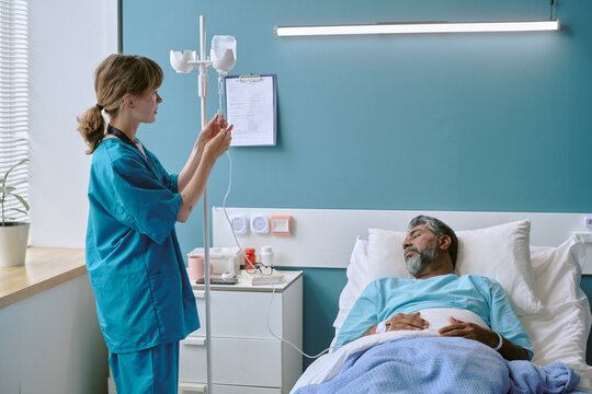 Young adult Caucasian female nurse adjusting IV drip for middle aged biracial man lying in hospital bed resting with eyes closed, medical equipment and clipboard visible in background