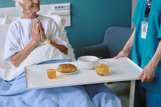 Senior Caucasian woman sitting in hospital bed smiling with hands clasped while female nurse serving breakfast tray with juice, bread roll, and bowl of cereal in medical setting
