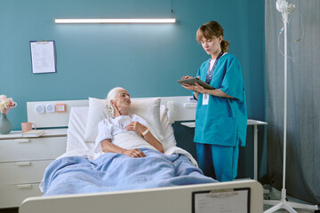 Senior Caucasian woman lying in hospital bed looking at young adult Caucasian female nurse standing beside bed holding digital tablet and writing medical notes