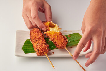 Crispy deep fried cheese sticks served on white plate with banana leaf garnish, showcasing gooey melted cheese filling as hands break one apart