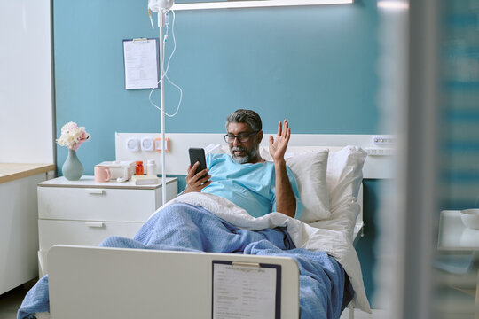 Middle aged biracial man lying in hospital bed waving and smiling while holding smartphone, engaging in video call with medical equipment and clipboard visible in background - Powered by Adobe