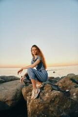 Lifestyle woman in modern boho-western denim street style sits on rocks by the sea at sunset in warm film color tones, capturing a relaxed and natural mood.