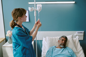 Caucasian young adult female nurse adjusting intravenous drip for middle aged biracial man lying in hospital bed with eyes closed, medical equipment visible in clinical setting