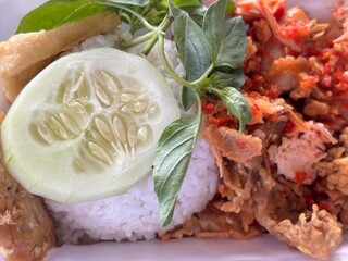 Traditional Indonesian spicy crushed fried chicken served with rice, cucumber, tempeh, and basil leaves. Street food concept with local flavor.