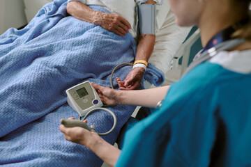 Middle aged Caucasian female nurse measuring blood pressure of senior Caucasian man lying in hospital bed, nurse holding digital blood pressure monitor, patient arm extended
