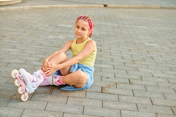 Thoughtful young girl in yellow tank top and striped shorts sitting on pavement, fixing her roller...