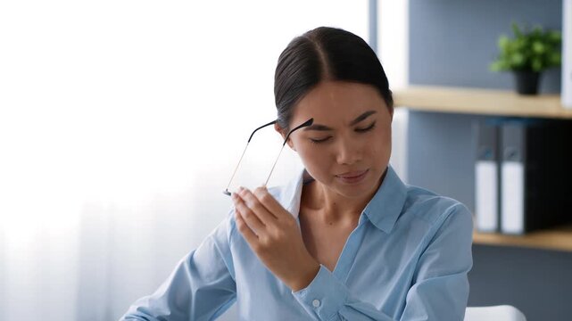 Young Asian woman in a light blue shirt works diligently at her desk in a modern office, adjusting her glasses while focusing on important tasks. Exhausted lady feeling sick