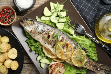 Tasty roasted seabass fish served with vegetables and lemon on wooden table, flat lay. Homemade seafood dish
