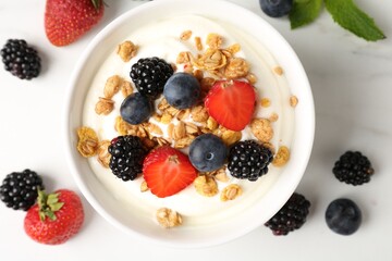 Tasty yogurt with granola and berries in bowl on white marble table, flat lay
