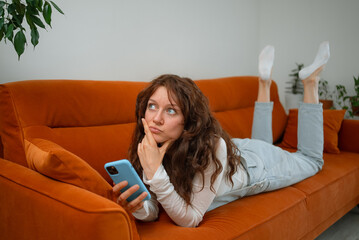 Thoughtful young woman with curly hair using mobile phone lying on comfortable orange sofa at home, thinking and looking away, holding smartphone and touching her chin