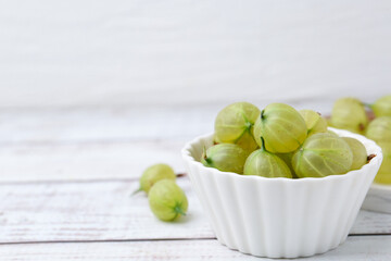 Fresh green gooseberries in bowl on white wooden table, closeup. Space for text