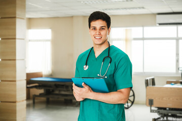 Smiling young male nurse or doctor in green scrubs holding a clipboard in a hospital room, representing professional healthcare, patient service, and modern medical support with a positive attitude.