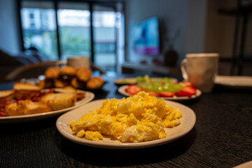 A plate of scrambled eggs sits on a table with other food. The table is set for breakfast, and there are cups and bowls nearby. Scene is casual and inviting