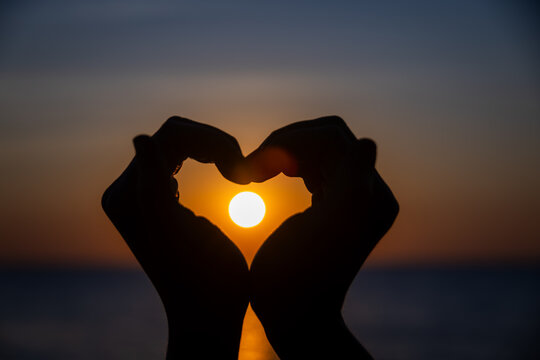 Silhouetted hands forming a heart shape perfectly framing the glowing sun as it sets over the ocean