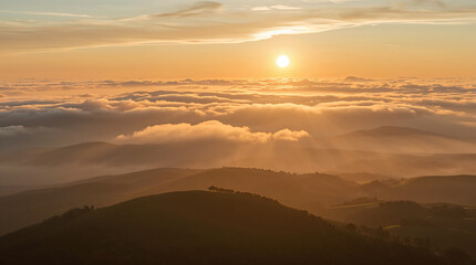 Golden sunset illuminates rolling hills shrouded in mist, with a sea of clouds below and sunbeams piercing through the atmospheric haze.