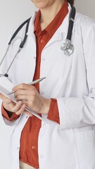 Close up of woman doctor wearing stethoscope and white coat over brick red blouse is standing and...