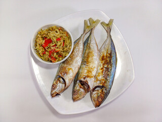 Three freshly fried fish arranged on a white plate, accompanied by a small bowl of spicy sambal, presented against a white background. Related to traditional Indonesian cuisine, authentic seafood dish