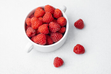 Raspberries in white bowl with scattered berries on light surface for organic food or vegan diet concept