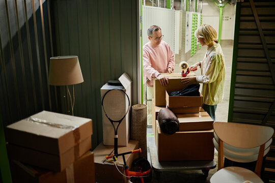 Middle aged Caucasian man and woman packing cardboard boxes together in storage facility, organizing belongings with various household items and sports equipment visible