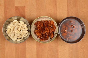 A wooden surface features three bowls: peeled almonds on the left, brown almond skins in the middle, and unpeeled almonds soaking in water on the right