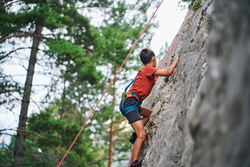 Fototapeta premium Young boy climbing rock wall with rope in forest environment