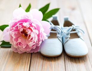 A delicate pair of light blue baby booties resting next to a blooming pink peony flower on a wooden table.