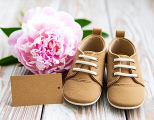 A close-up of a pair of soft brown baby shoes next to a blooming pink peony and a blank card on a wooden table.