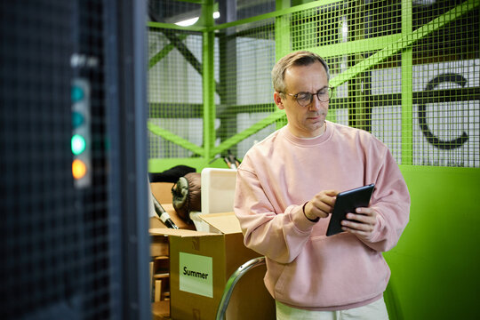 Caucasian middle aged man wearing glasses using digital tablet while standing in storage facility near cardboard boxes labeled with seasons, focusing on organizing belongings
