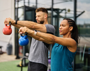 Portrait of a young black woman and white man coach or trainer exercising in a gym, lifting weights, kettlebell equipment, healthy lifestyle and strength exercise at fitness club concepts