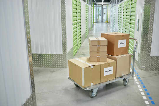 Stacked cardboard boxes labeled Summer and Winter sitting on metal cart positioned in corridor of self storage facility with green doors, concrete floor and industrial lighting visible