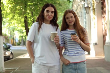 Portrait of beautiful mother and her teenage daughter with paper cups on city street