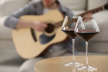 Glasses with red wine on table and man playing guitar indoors, selective focus