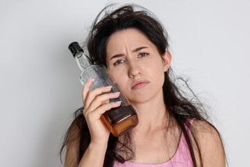 Woman with bottle of whiskey suffering from hangover on light grey background
