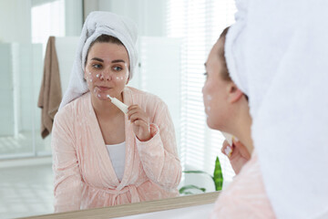 Woman applying cream onto face near mirror indoors. Acne treatment
