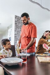 Father preparing breakfast while standing at the kitchen table with his family. Child watches with interest as family enjoys a cozy morning meal together.