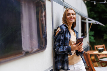 Happy woman standing near camper van, using smartphone on camping trip in countryside, copy space.