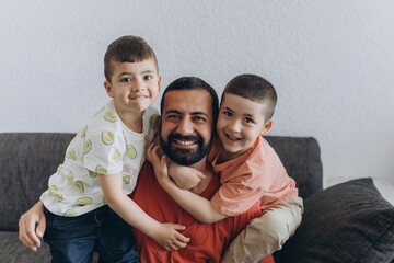 Happy father hugging two smiling sons on a couch. Family bonding moment with kids embracing their dad with love and laughter. Warm, joyful scene of fatherhood and parenting