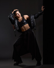 Beautiful sexy brunette girl in black leather zippered bra, leather jacket, high boots and black pants posing in studio on grey background