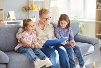 Smiling grandmother sitting on gray sofa and reading storybook to two children of different age. Happy family enjoying leisure time and storytelling in living room at home. Child development concept.